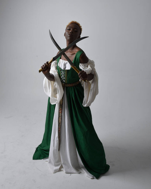 Full length portrait of pretty African woman wearing long green medieval fantasy gown holding  sword, standing action pose on a light grey studio background. - Photo, Image