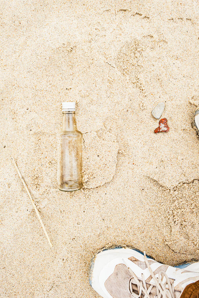 Una vera bottiglia di vetro trovata sulla spiaggia sulla sabbia accanto a un piede in una sneaker bianca e rocce marine  - Foto, immagini