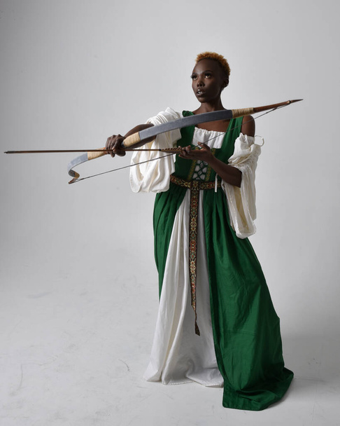 Full length portrait of pretty African woman wearing long green medieval fantasy gown holding bow and arrow, standing action pose on a light grey studio background. - Photo, Image