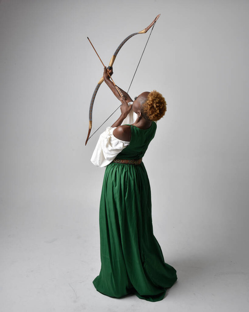 Pełna długość portret pięknej afrykańskiej kobiety noszącej długą zieloną średniowieczną suknię fantasy trzymającej łuk i strzałę, stojącej pozycji akcji na jasnoszarym tle studio. - Zdjęcie, obraz