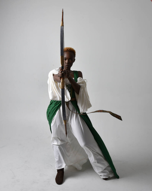 Teljes hosszúságú portré csinos afrikai nő visel hosszú zöld középkori fantasy ruha tartó íj és nyíl, álló fellépés pózol egy világosszürke stúdió háttér. - Fotó, kép