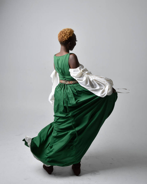 Полнометражный портрет красивой африканской женщины в длинном зеленом средневековом фэнтези-платье, стоящей лицом назад на светло-сером фоне студии. - Фото, изображение