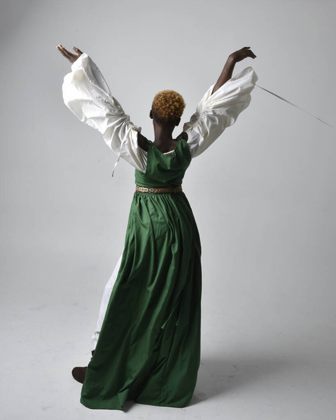 Ritratto a figura intera di bella donna africana che indossa lungo abito fantasia verde medievale, in piedi posa rivolta all'indietro su uno sfondo grigio chiaro studio. - Foto, immagini