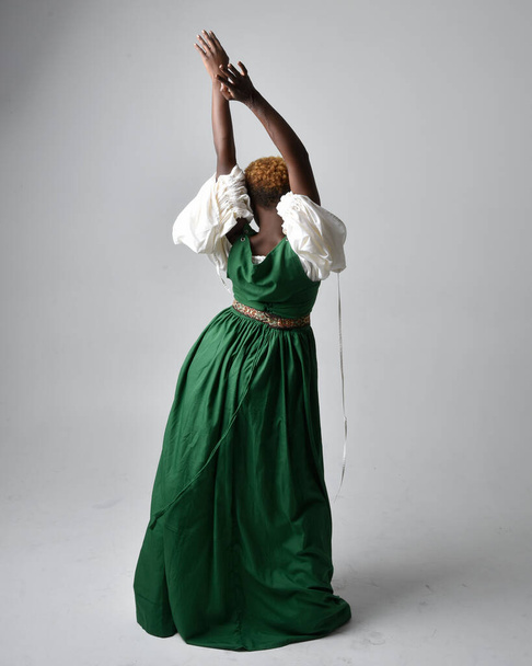 Πλήρες πορτρέτο της όμορφης Αφρικανής γυναίκας που φοράει μακρύ πράσινο μεσαιωνικό φόρεμα φαντασίας, στέκεται ποζάροντας προς τα πίσω σε ένα ανοιχτό γκρι φόντο στούντιο. - Φωτογραφία, εικόνα
