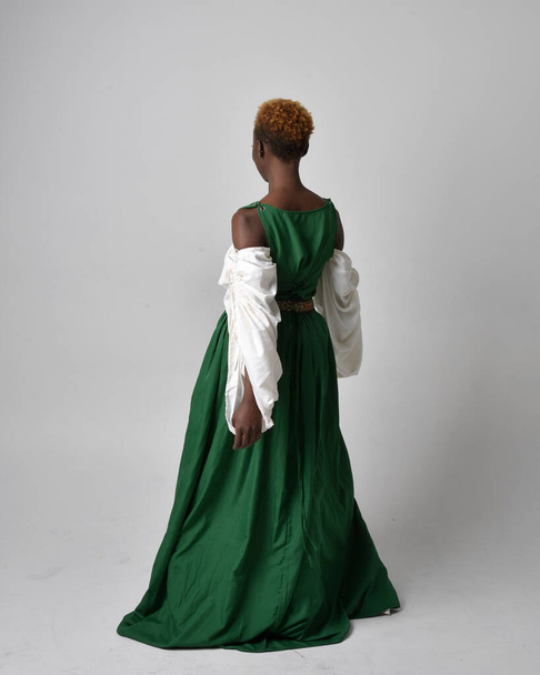 Full length portrait of pretty African woman wearing long green medieval fantasy gown, standing pose facing backwards on a light grey studio background. - Фото, изображение