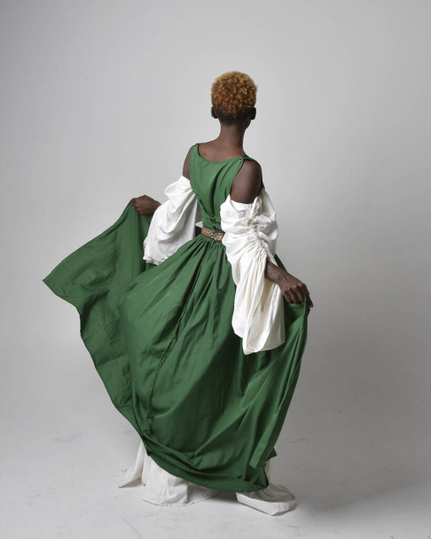 Full length portrait of pretty African woman wearing long green medieval fantasy gown, standing pose facing backwards on a light grey studio background. - Photo, Image