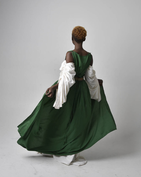 Full length portrait of pretty African woman wearing long green medieval fantasy gown, standing pose facing backwards on a light grey studio background. - Φωτογραφία, εικόνα
