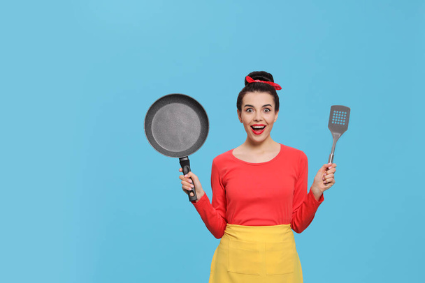 Emotional housewife with frying pan and spatula on light blue background. Space for text - Foto, Bild