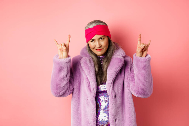 Funny and cool asian senior woman showing rock n roll gesture, looking sassy, standing over pink background - Foto, immagini