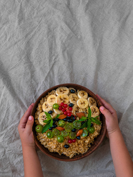 Wholegrain Oatmeal porridge bowl with fresh berries fruit nut mint child hands flat lay. Healthy food oat meal morning breakfast top view. Whole grain cereal gooseberry currant banana linen tablecloth - 写真・画像