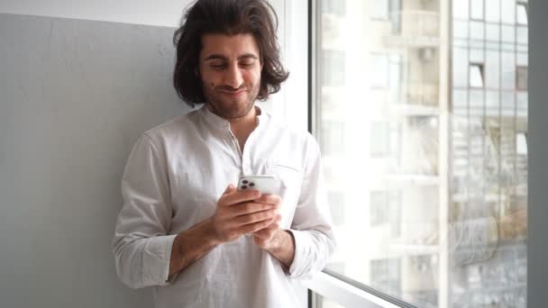 Удовлетворенный взрослый турецкий мужчина смотрит на свой мобильный, стоящий возле окна в помещении. - Кадры, видео