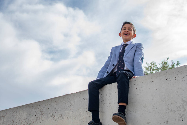 Laughing schoolboy in formal suit with classic smart white shirt and tie looking at the camera while sitting on top of a concrete wall - Fotoğraf, Görsel