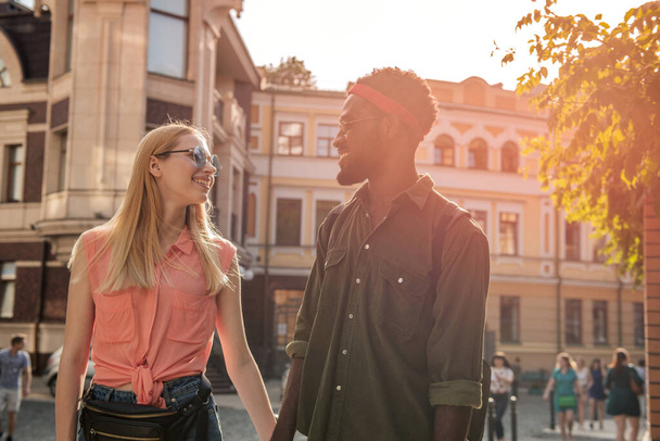 Ein junges, fröhliches Paar, in dem ein dunkelhäutiger Mann und eine blonde Frau lächelnd einander an den Händen halten, während sie eine Straße entlang gehen - Foto, Bild