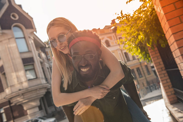 Young cheerful couple where a black man and a blonde woman are smiling. Girl sitting on the back of a man hugs his neck while walking around the city - Foto, imagen