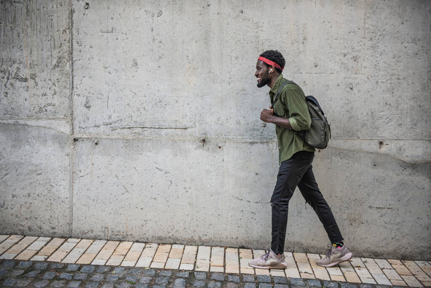 Ein junger schwarzer Mann läuft auf dem Bürgersteig, während er mit einem Rucksack in der Nähe einer großen Mauer durch die Straßen einer Brücke läuft - Foto, Bild