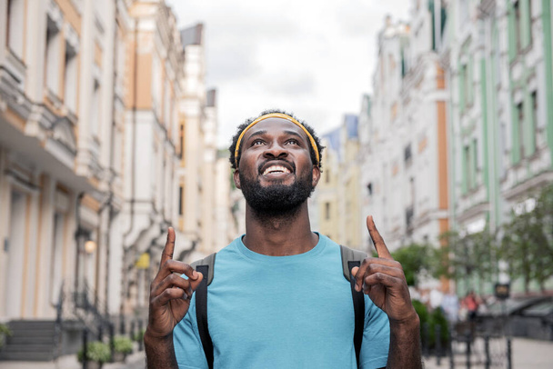 Młody czarny człowiek raduje się podnosząc palce podczas spaceru ulicami mostu z plecakiem. - Zdjęcie, obraz