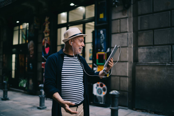 Elderly Caucasian male tourist in electronic headphones making international video call in roaming using modern touch pad and 4g internet, happy man 60s reading network news on gadget website - Foto, immagini