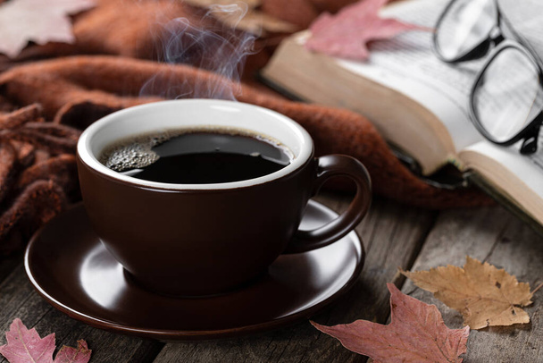 Closeup of a steaming hot cup of coffee with book, blanket and autumn leaves on a rustic wooden table - Fotó, kép