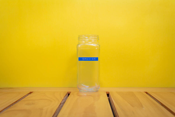 Frasco de vidrio vacío, con una etiqueta azul claro que dice "Ahorros", sobre un fondo amarillo - Foto, imagen