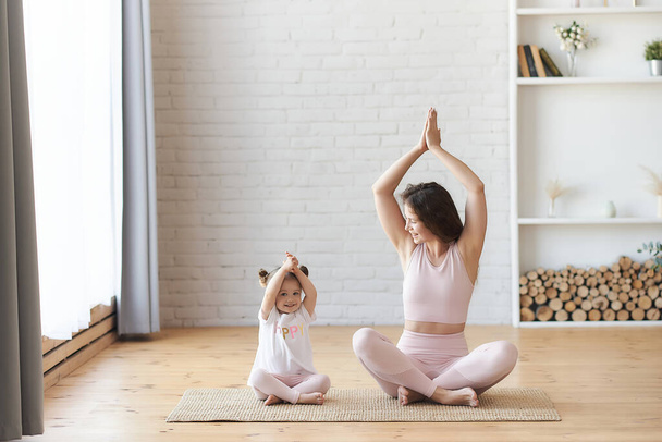 Family mother and child daughter are engaged in fitness, yoga, exercise at home. Smiling young curly woman looking at her small kids girl sitting on the carpet floor in lotus pose namaste hands up - Фото, изображение