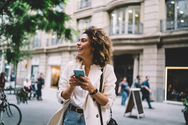 Cheerful travel blogger rejoicing at touristic street using cellular device for roaming networking during solo vacations, funny hipster girl with mobile technology laughing in international city - Foto, Imagen
