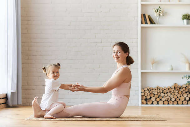 Mooie jonge vrouw en haar charmante dochtertje glimlachen terwijl ze thuis yoga doen, passen gezinsyoga toe in de huiskamer en herstellen de gezondheid na de bevalling - Foto, afbeelding
