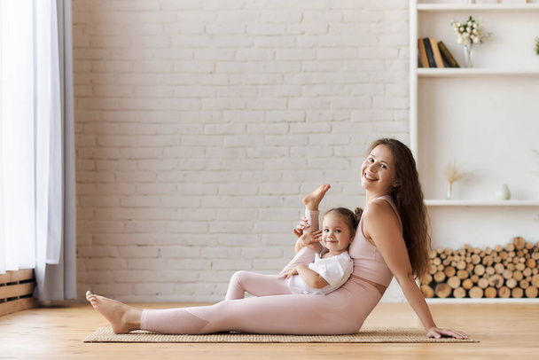 Jonge vrouw met lang krullend haar in licht roze outfit zit op de vloer kijken en glimlachen naar de camera met kleine baby meisje zitten op haar knieën en tilt haar been omhoog voor spierflexibiliteit. - Foto, afbeelding
