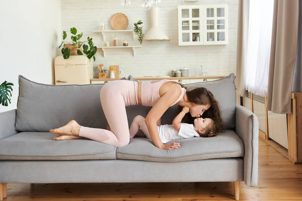 Happy young mother in light pink sportswear with curly long hair holding and kissing cute daughter lying on couch at home. Close up relaxing parent having fun free time and playing with child. - Foto, Imagem