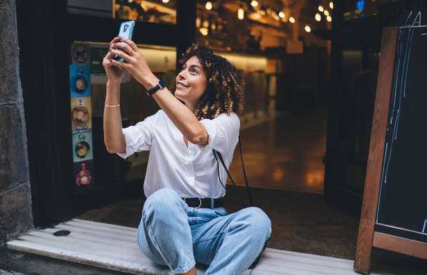 Happy Caucaisan hipster girl photographing city while resting near doorway using cellphone device for web networking, pretty female travel blogger connecting to online streams for shooting vlog - 写真・画像