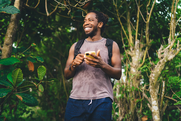 Happy African American male tourist in casual clothes with backpack smiling and browsing mobile phone while standing near green tropical plants and looking away - Foto, Imagen