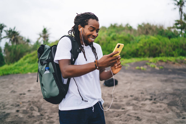 Excited black male tourist in casual clothes standing on sand near tropical green trees and chatting via smartphone using earphones on blurred background - Zdjęcie, obraz