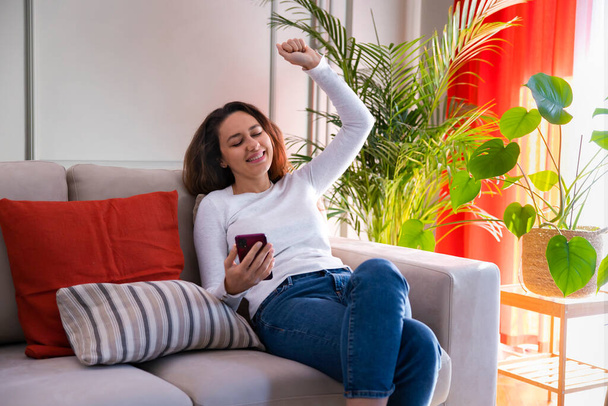 Happy young woman getting good news notification via mobile phone, making yes gesture, celebrating success sitting on couch - Fotografie, Obrázek