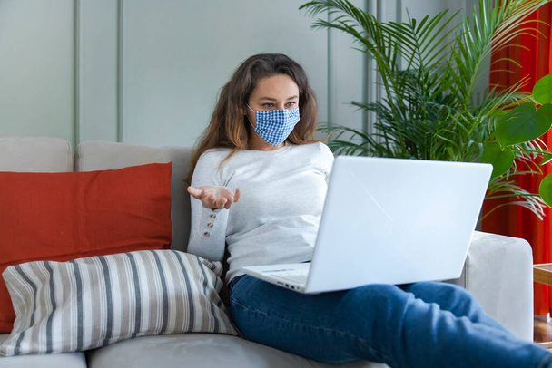 Young woman with face protective mask chatting online with laptop at home - Foto, afbeelding