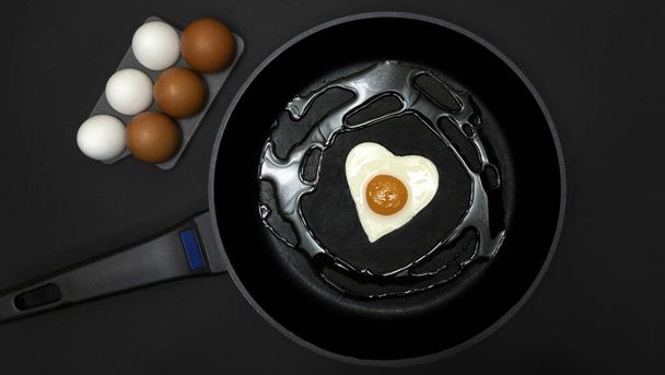 Una sartén con un huevo frito en forma de corazón y al lado media docena de huevos blancos y marrones, todo sobre un fondo negro, cocina saludable y concepto de colesterol - Foto, imagen