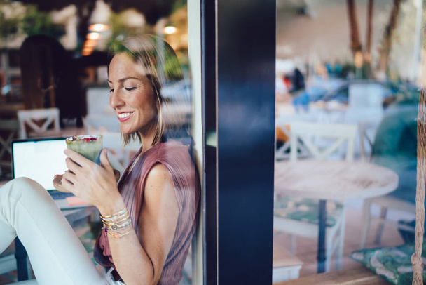 durch ein Glas positiver Frauen in stilvollem Outfit genießen eine Tasse leckeres Getränk und sitzen in einem modernen Café mit Laptop - Foto, Bild