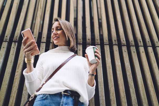 снизу счастливая женщина в повседневной одежде читает новости на смартфоне и пьет кофе на вынос во время перерыва на улице - Фото, изображение