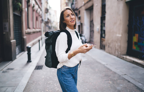 Caucasian tourist with backpack using smartphone application for searching location during sightseeing excursion, beautiful woman with cellphone technology exploring city quarters and streets - Foto, Imagem