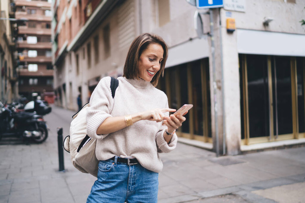 Cheerful stylish lady in casual clothes and backpack writing SMS on smartphone and smiling while standing on sidewalk in downtown - Foto, Imagem