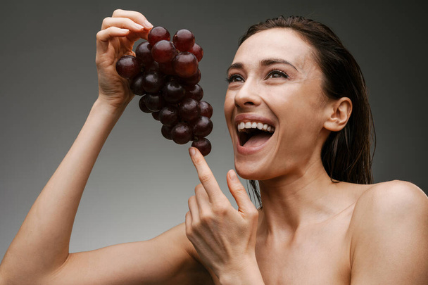 White shirtless woman laughing while posing with grapes isolated over grey background - Foto, Bild