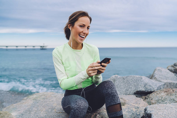 Portrait of joyful jogger in sportive tracksuit resting at seashore stones enjoying motivation music podcast, happy fit girl listening positive audio songs downloaded to smartphone technology - Zdjęcie, obraz