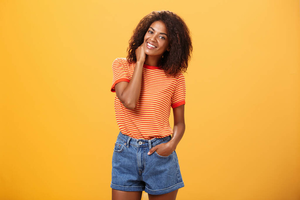 Стильная афро-американская стройная женщина с вьющимися средними волосами, держась за руку в джинсовых шортах, наклоняя голову, потирая шею, улыбаясь дружелюбно чувствуя себя неуютно или стесняясь в новой компании над оранжевой стеной - Фото, изображение