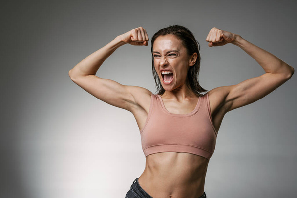 White woman wearing brassiere screaming while showing her biceps isolated over grey background - Photo, image