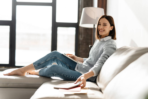 Young smiling woman using tablet computer while sitting on sofa at home - Фото, изображение