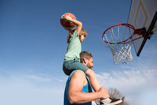 Father and son playing basketball. Happy father holding his little son on shoulders, helping him to score a basket on a basketball playground. Copy space. - 写真・画像