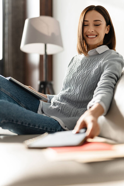 Молода усміхнена жінка використовує планшетний комп'ютер, сидячи на дивані вдома
 - Фото, зображення