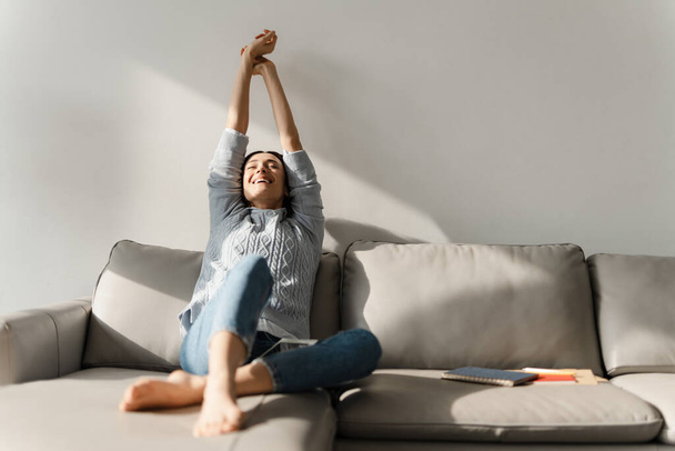 Young smiling woman stretching her body while sitting on sofa at home - Foto, Imagem