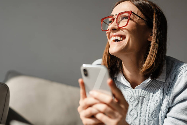 Young laughing woman in eyeglasses using cellphone while sitting on sofa at home - Fotoğraf, Görsel