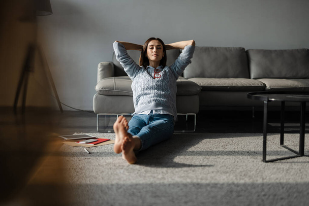 Giovane donna bianca seduta sul pavimento con le mani strette dietro il collo a casa - Foto, immagini