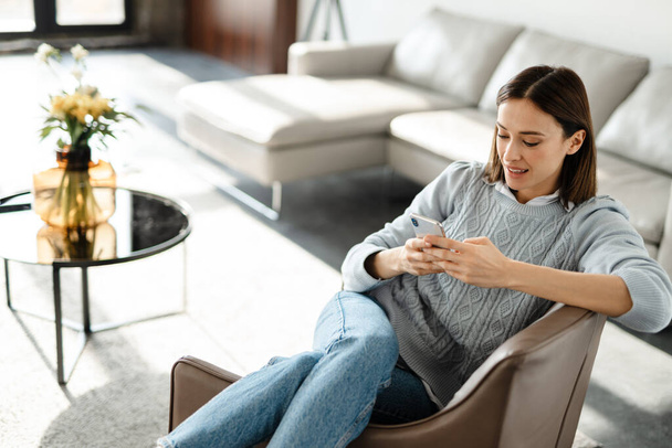 Young white woman using cellphone while sitting in armchair at home room - Fotoğraf, Görsel