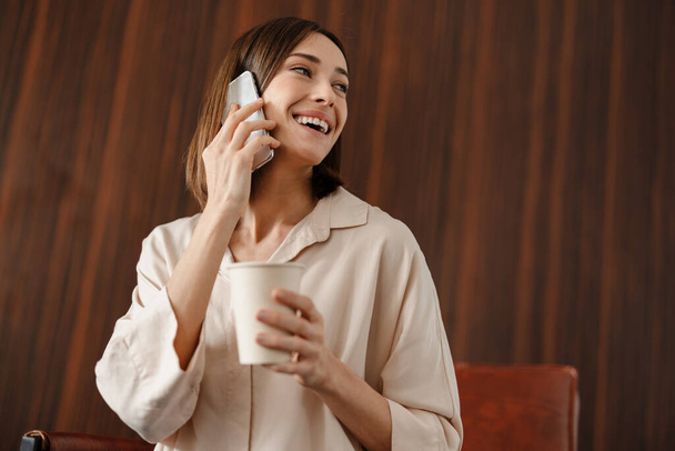 Smiling woman drinking coffee and talking on cellphone while sitting in armchair indoors - Photo, image